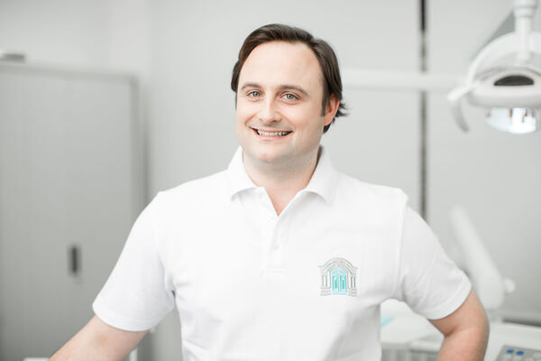 Ein lächelnder Zahnarzt in weißem Kittel steht in moderner, heller Zahnarztpraxis.