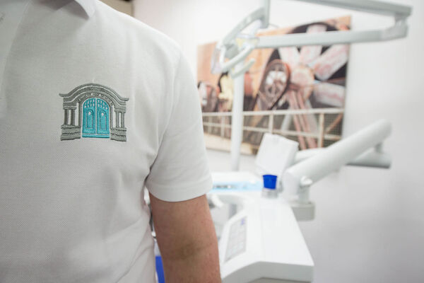 Mitarbeiter im weißen Poloshirt mit Logo steht in moderner Zahnarztpraxis am Behandlungsstuhl.